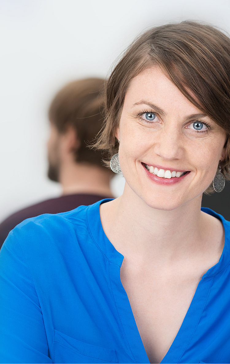 Eine lächelnde Frau mit kurzen, braunen Haaren trägt ein blaues Hemd. Sie sitzt in einem modernen Büro und schaut direkt in die Kamera. Im Hintergrund sind unscharf zwei weitere Personen bei der Arbeit zu sehen.
